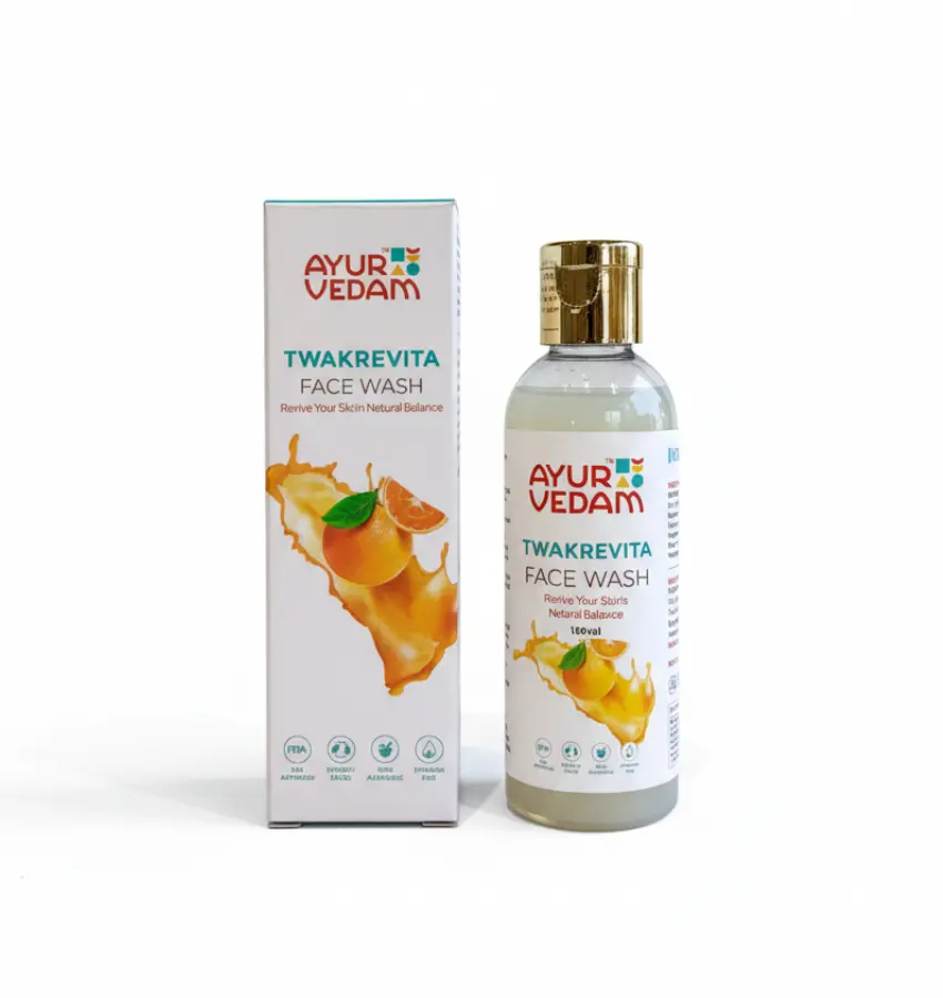 A 100ml bottle and matching box of Ayurvedam Twakrevita Face Wash. The packaging is white with a gold flip-top cap and features an illustration of a fresh orange splash. The label states "Revive Your Skin's Natural Balance" and displays icons for FDA approval and sulfate-free formula.