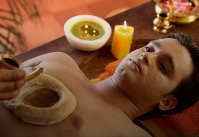 A person at Ayurvedam Store receiving a relaxing Ayurvedic oil treatment named hridya basti, with a lit candle and bowl of oil nearby.