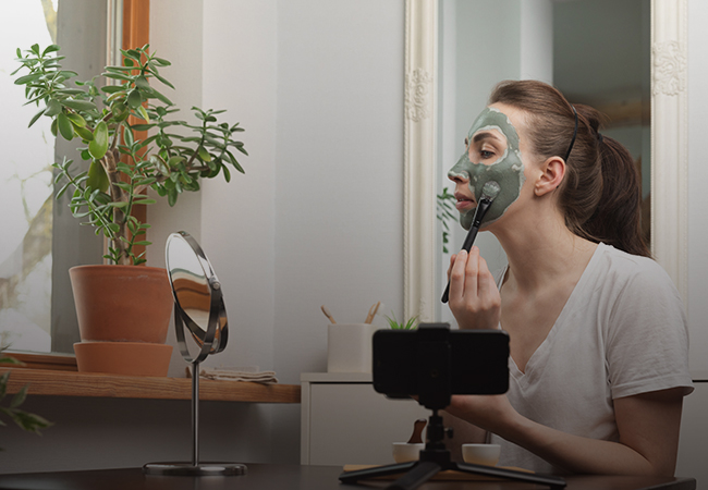 Person applying ayurvedam's instant glow facial cream in front of a round mirror, in a room lit by natural light from a window near a potted plant.