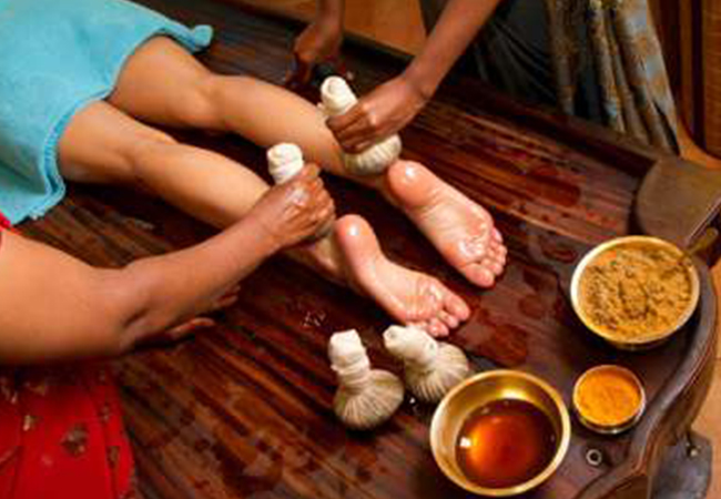 A person is getting a Leg Potli Massage with herbal bundles at Ayurvedam Store. Bowls of natural ingredients are nearby.