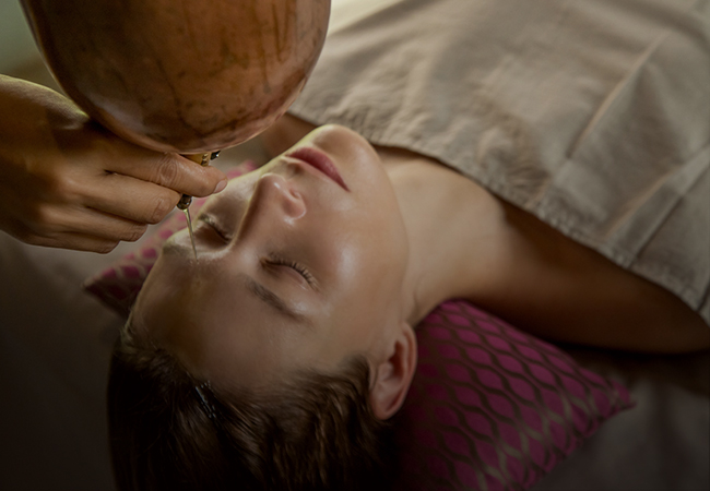 A person at Ayurvedam Store receiving a traditional Ayurvedic treatment with herbal oil poured from a clay pot onto their forehead.