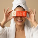 A girl in a white robe and towel, holding Kumkumadi Elixir Oil skincare product in both hands against a beige background