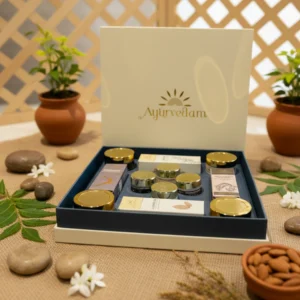 A luxurious Ayurvedic gift box labeled "Ayurvedam," containing various skincare or wellness products in jars and boxes with gold and silver lids. The box is surrounded by natural elements like almonds, jasmine flowers, neem leaves, smooth stones, and potted plants, all arranged on a burlap surface for an earthy and calming aesthetic.